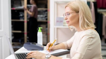 Experienced bookkeeper reviewing retail sales records on a laptop inside a clothing store office while a staff member checks the inventory
