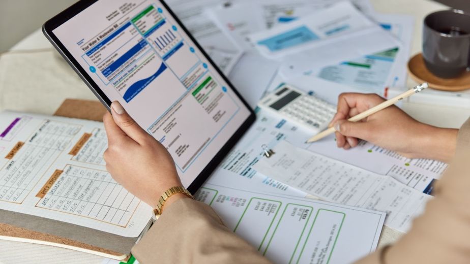 A bookkeeper cross-checking financial documents with tablet data while recording notes 