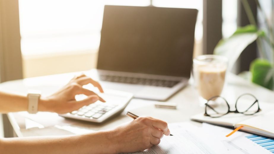 A bookkeeper recording transactions on paper using a calculator, with a laptop, coffee cup, and eyeglasses on the desk