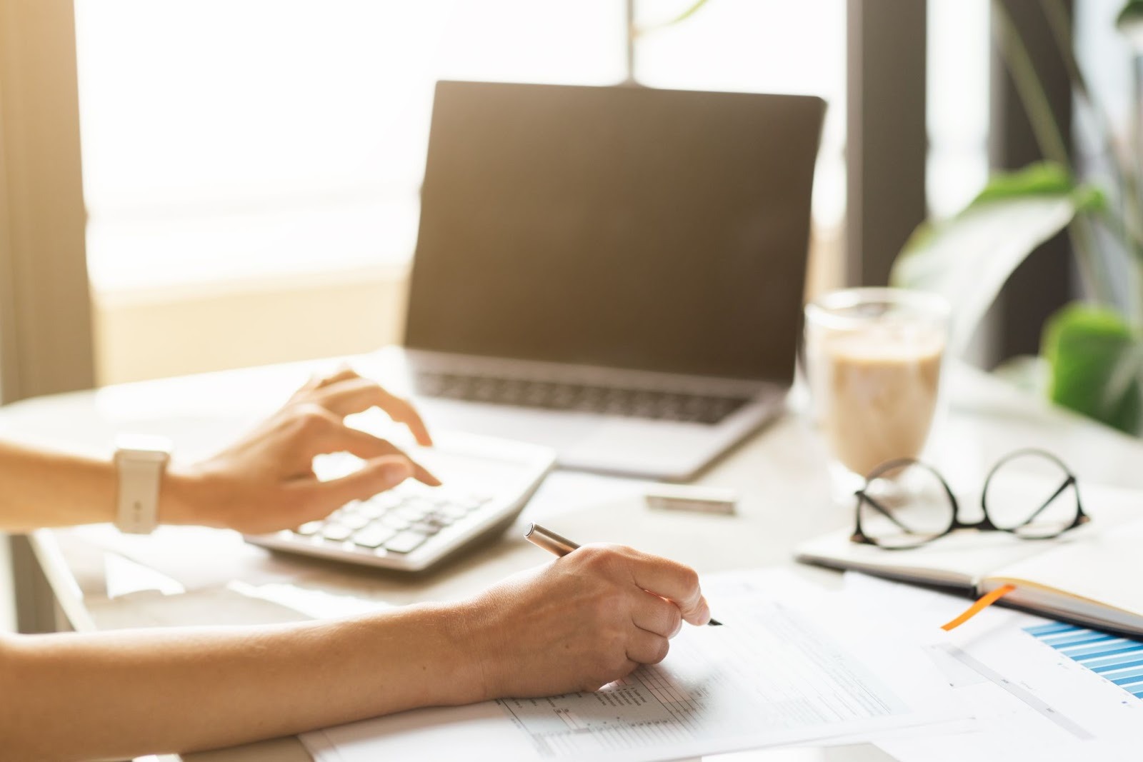 A bookkeeper recording transactions on paper using a calculator, with a laptop, coffee cup, and eyeglasses on the desk
